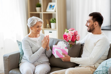 family, mother's day and birthday concept - smiling adult son giving present and flowers to his senior mother at home