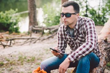 Young man sit on truck in forest and using mobile phone. man traveler sits on large fallen tree, holding phone in hand. theme hiking nature travel. Outdoor activities hiking. Traveling and adventure