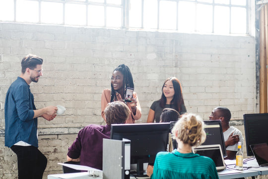 Cheerful businesswoman showing mobile phone to team while looking at male colleague in office
