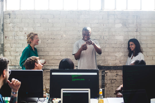 Smiling young colleagues looking at male programmer using smart phone against wall in office