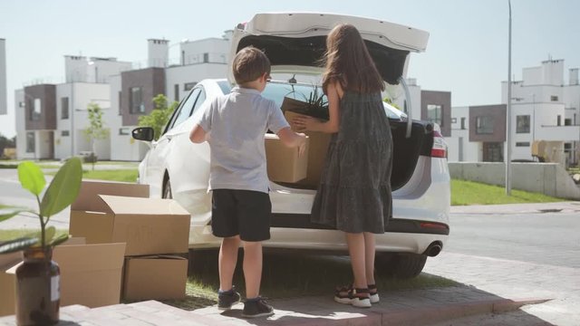 Back view of happy children loading boxes into a car trunk and sitting inside. Young adults moving into a new house on a sunny summer day.