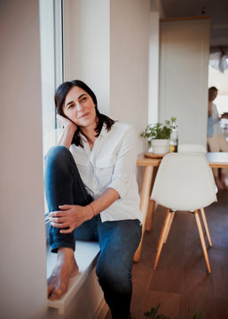 Mature woman sitting on windowsill in the kitchen, daydreaming