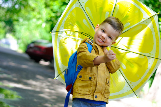 A little cute boy in a yellow raincoat stands in a puddle and holds an umbrella with a lemon coloring.	