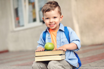 Fototapeta premium A small three year old preschooler with a backpack on his back and with books sits on the stairs and shows his green apple for a snack.