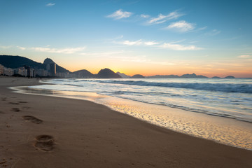 Footsteps along the ocean waves on Copacabana beach in Rio de Janeiro with the Sugarloaf mountain in the background at early morning orange and blue sunrise