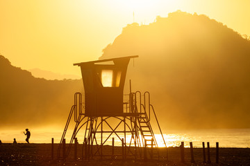 Silhouette of an old lifeguard tower against a glowing haze on the beach of Copacabana in Rio de Janeiro backlit by intense golden hour sunlight with a mountain in the background