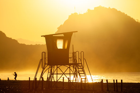 Silhouette of an old lifeguard tower against a glowing haze on the beach of Copacabana in Rio de Janeiro backlit by intense golden hour sunlight with a mountain in the background