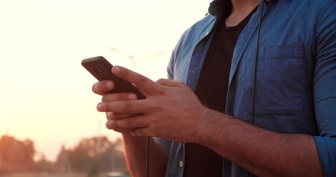 Closeup of Man’s Hands browsing his Smartphone and texting Message in New App. Handsome Bearded Man listening to Music in Headphones and smiles. Beautiful Sunset on the Background. Games. Emotions.