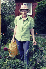 Portrait of elderly woman in a hat stands near the beds in her garden with a watering can.