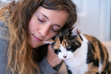 Closeup portrait of happy smiling young woman bonding with calico cat pet, bumping rubbing bunting heads, showing affection