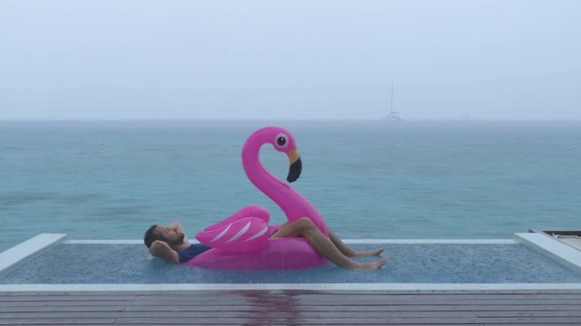 Vacation rain - funny video of holidays getaway travel raining away under heavy rainfall with relaxing and happy man smiling on flamingo float in luxury pool with funny face expression.