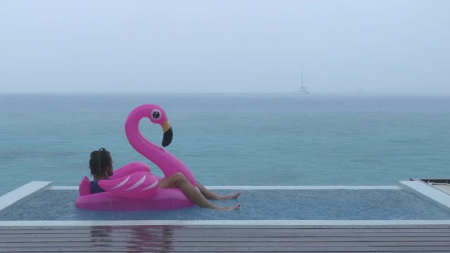 Vacation rain - funny video of holidays getaway travel raining away under heavy rainfall with sad and unhappy man on flamingo float in luxury pool with funny face expression.