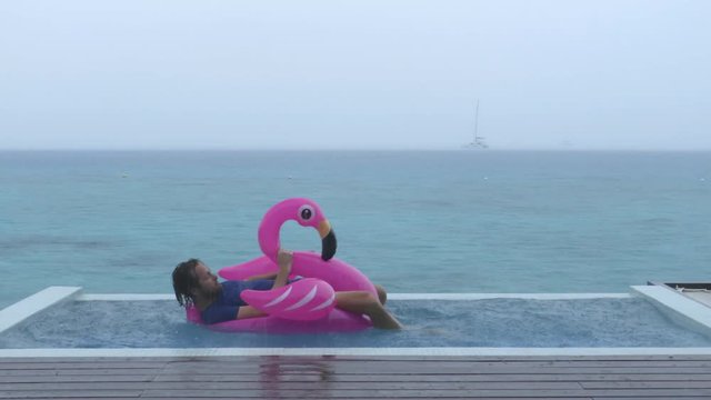 Funny fail video in vacation rain of holidays getaway travel raining away under heavy rainfall with man falling while trying to get out of flamingo float in luxury pool with funny face.