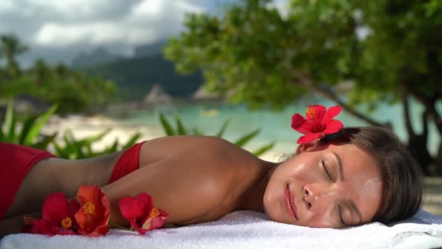 Spa Woman relaxing getting massage at hotel resort. Beauty girl lying down sleeping on towel, hibicus flowers outside. Serene ethnic woman relaxing. Mixed race Asian / Caucasian model outdoor