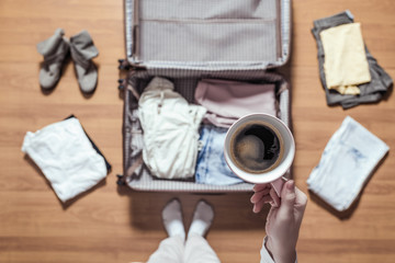 Top view of woman hands packing a luggage for a new journey and drinking coffee from the pink cup .