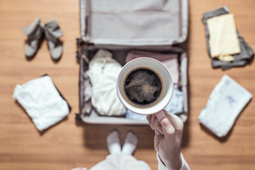 Top view of woman hands packing a luggage for a new journey and drinking coffee from the pink cup .