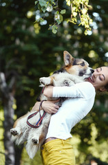 Smiling young woman laughing, holding cute dog Welsh Corgi in a park outdoors. Beautiful happy female in white shirt playing with her Welsh Corgi Pembroke doggy outside. Concept friendship with dog