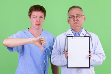 Stressed mature Japanese man doctor with Scandinavian man patient showing clipboard and giving thumbs down