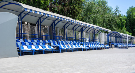 Football, stadium, area for spectators and coach. Empty blue and white plastic armchairs under a canopy. Place for text.