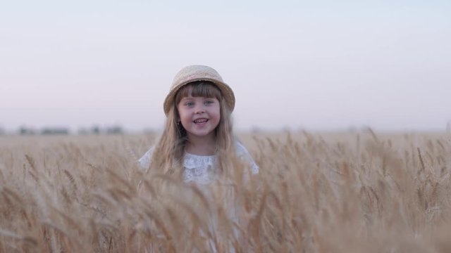 happy village childhood, little cute girl with straw hat having fun and laughing spins in reaped grain wheat spikes at harvest season against sky