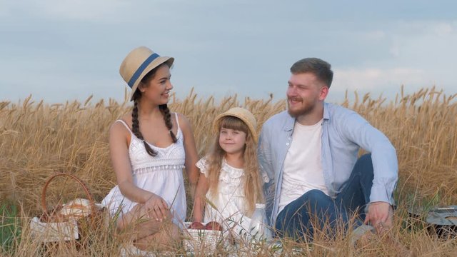 happy young family, cheerful parents with little cute child girl hug and look at each other smiling and sitting at outing in grain harvest autumn barley field shining by sun