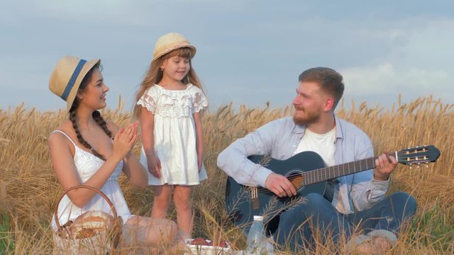 cheerful family outings, father plays string musical instrument while mom claps her hands and little sweet daughter dances in yield seasonal bread field