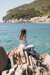 The girl sits on a rocky seashore and admires a beautiful view of the coastal city of Petrovac in Montenegro.