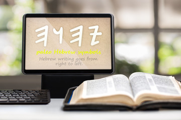 A tablet showing the paleo Hebrew symbols for the name of God on a desk with a keyboard, opened holy bible and cross in background.