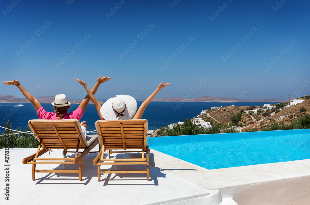 Wall mural Urlaubskonzept: glückliches Paar auf Sonnenliegen am Pool streckt die Arme in die Luft und genießt die Aussicht auf das Meer