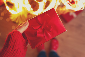 Woman hand holding red Christmas gift box wrapped red ribbon against background of Christmas tree...