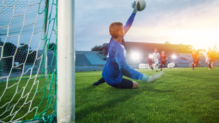 On Stadium Attacker Kicks the Ball and Scores the Goal, Soccer Goalkeeper Tries to Defend Goals Jumps and Catch the Ball. Shot with Warm Sunlight Flare in the Background.