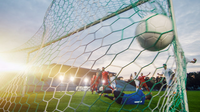 On Soccer Championship Goalkeeper Tries to Defend Goals but Jumps and Fails to Catch the Ball. Shot from Behind the Net with the Ball in it. Stadium Shot with Warm Sunlight Flare in the Background.