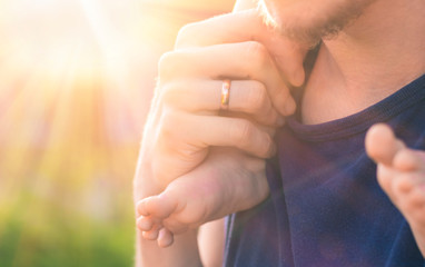 Man's hand with a wedding ring on his finger, holding the foot of his bare little child. Toddler is sitting at his father's neck on the background of soft sunlight and blurred green bokeh.