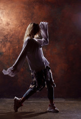 Fototapeta na wymiar Photo of young dancing blonde looking to side in torn jeans and sneakers on dark brown background