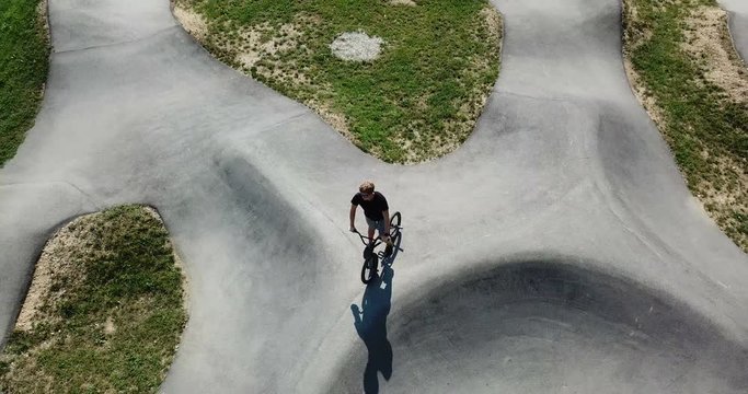 AERIAL 4K: Sport activity. Young male bmx rider casually dressed standing with bicycle on the concrete pump track on a sunny day. Camera top down, circling subject.