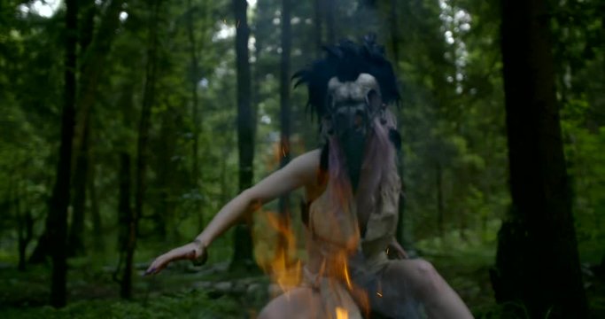 shamanic woman is performing ritual movements in forest near fire, wearing rags