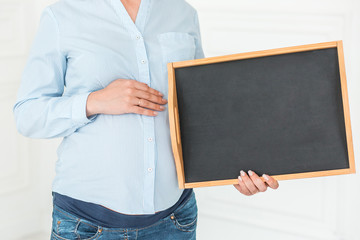 Pregnant woman is holding an empty blackboard. Close-up, copy space, indoors.