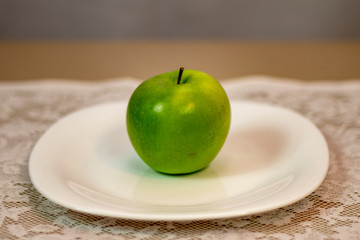 Green apple served on a white porcelain plate on a table covered with white lace towel.