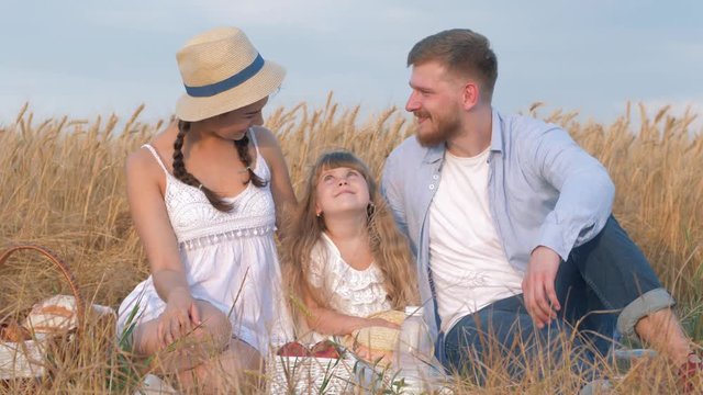 happy family portrait, young couple sits together close to their child girl daughter in open air outings in harvest field of matured grain wheat spikes against sky