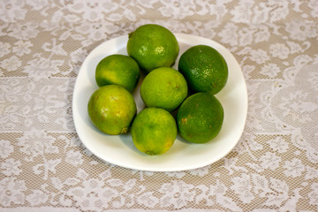 Green lemons served on white porcelain plate on a table covered with white lace towel.