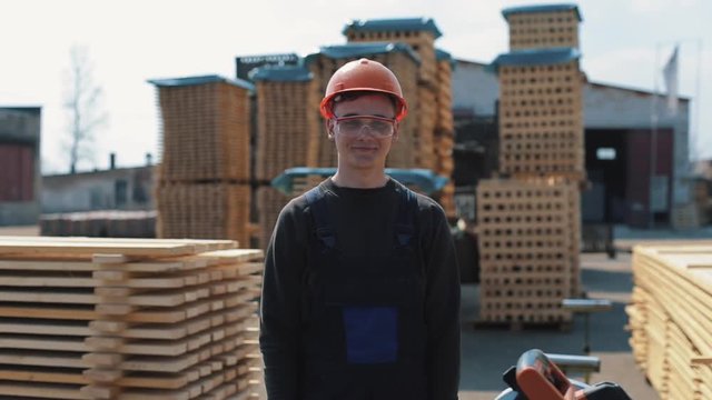 Industrial young worker wearing construction helmet and shows thumbs up. A lot of pallets on the background.