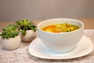  Soup of cooked vegetables with broth served in white bowl on white tablecloth tablecloth garnished with succulent flowers.