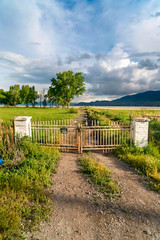 Dirt road with rusty metal gate on a vast grassy field in front of a lake