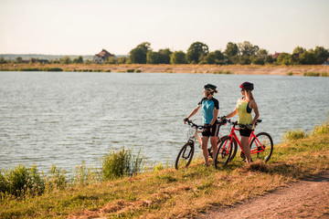 Fototapeta premium Two women cyclists with bikes standing and talking at the lake shore
