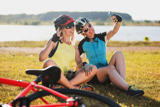 Two friends women wearing cycle gear sitting on grass and take selfie