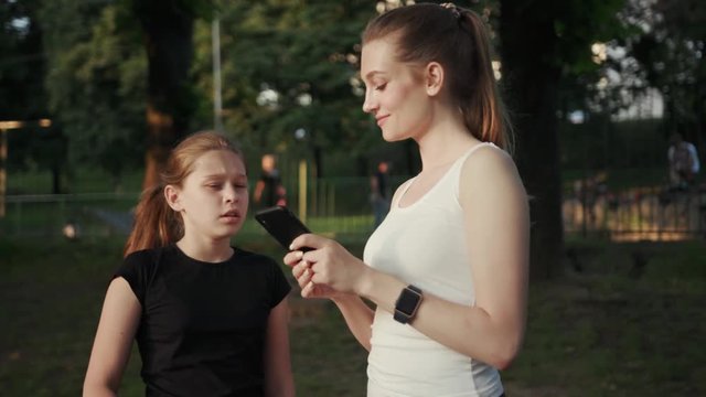 Nice-looking beauty fit sisters spend time in the city park watch videos on smartphone. Lovely sporty teen girl sneezing looking on screen standing with her sister in the street at sunlight.