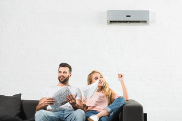 couple on couch with newspapers suffering from heat under air conditioner at home