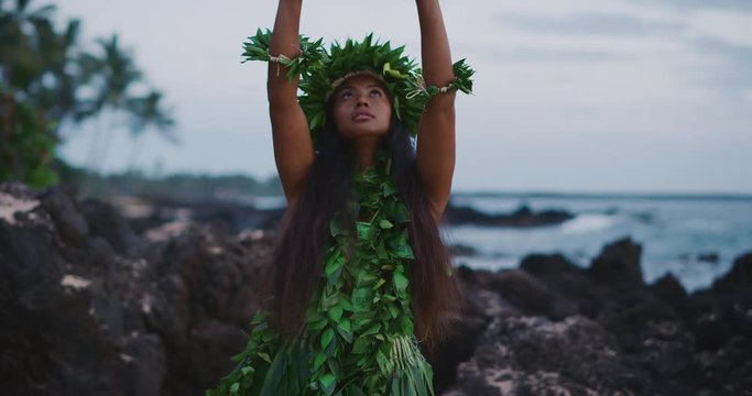 Traditional Hawaiian hula dancing at sunset in slow motion, woman performing Hawaiian hula with haku leis and ti leaf skirt with the ocean in the background