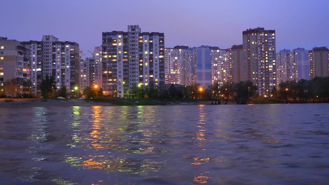 A complex of appartment buildings in the evening city near the large lake, with street lights reflecting in the water. Buildings in twilight near lake. Buildings and lights in appartments evening.