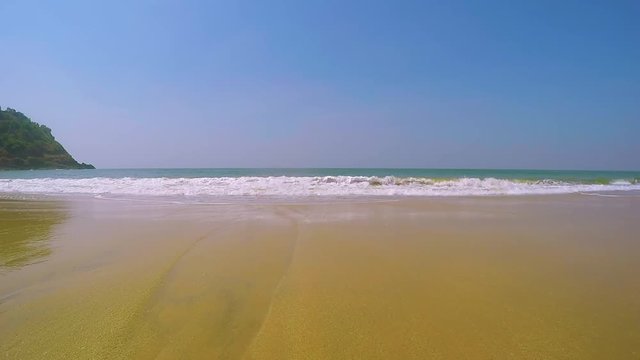 Small waves crush on the shore and move towards the camera.  Blue water with white foam, beach with yellow fine sand on a tropical sunny day